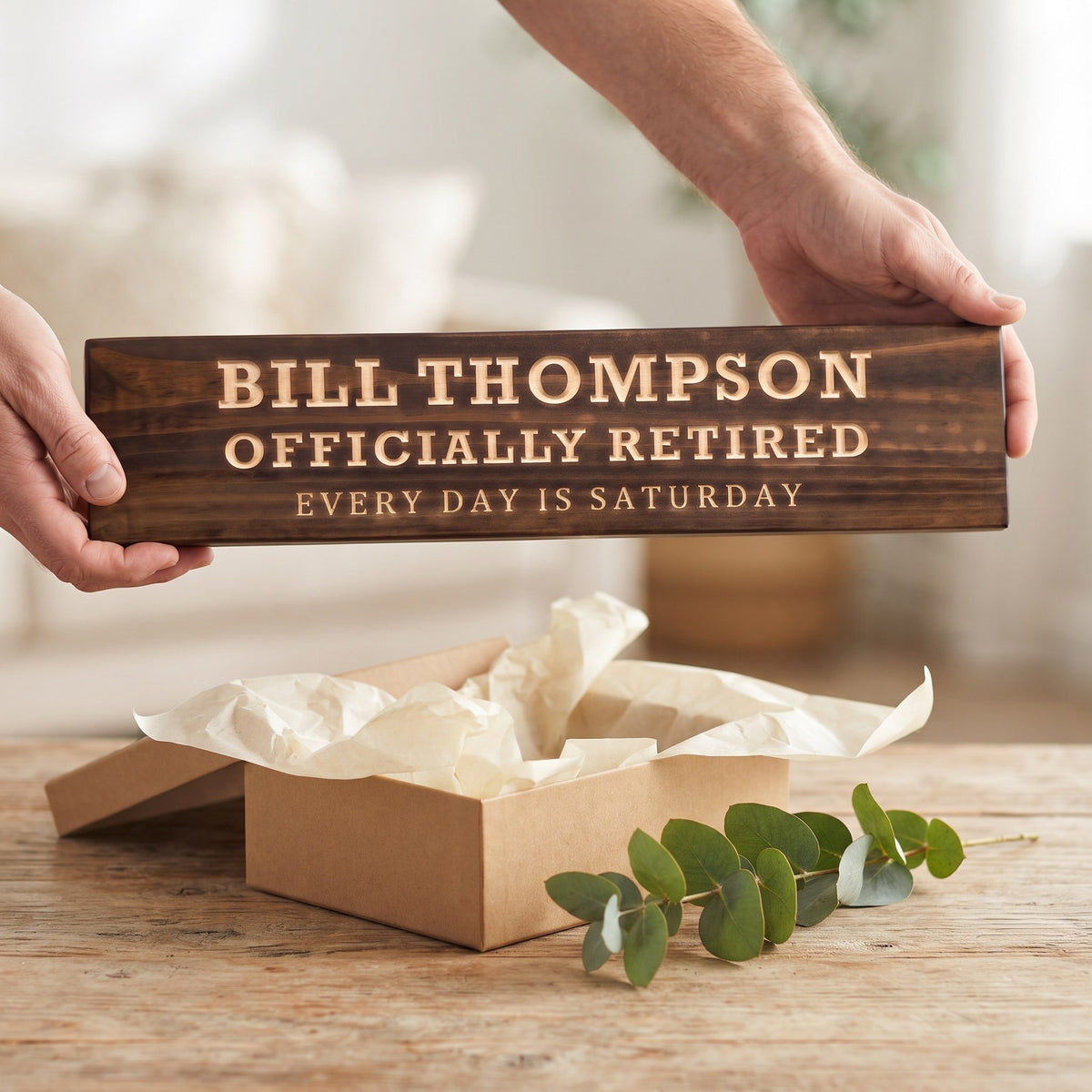 Hands holding a personalized wood sign that reads Bill Thompson over a kraft gift box with tissue paper and twine