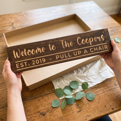 Hands holding a personalized wood sign that reads Welcome to The Coopers' over a kraft gift box with tissue paper and twine