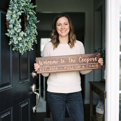 A smiling woman in a light sweater standing at her open front door holds a personalized wood sign that reads Welcome to The Coopers'