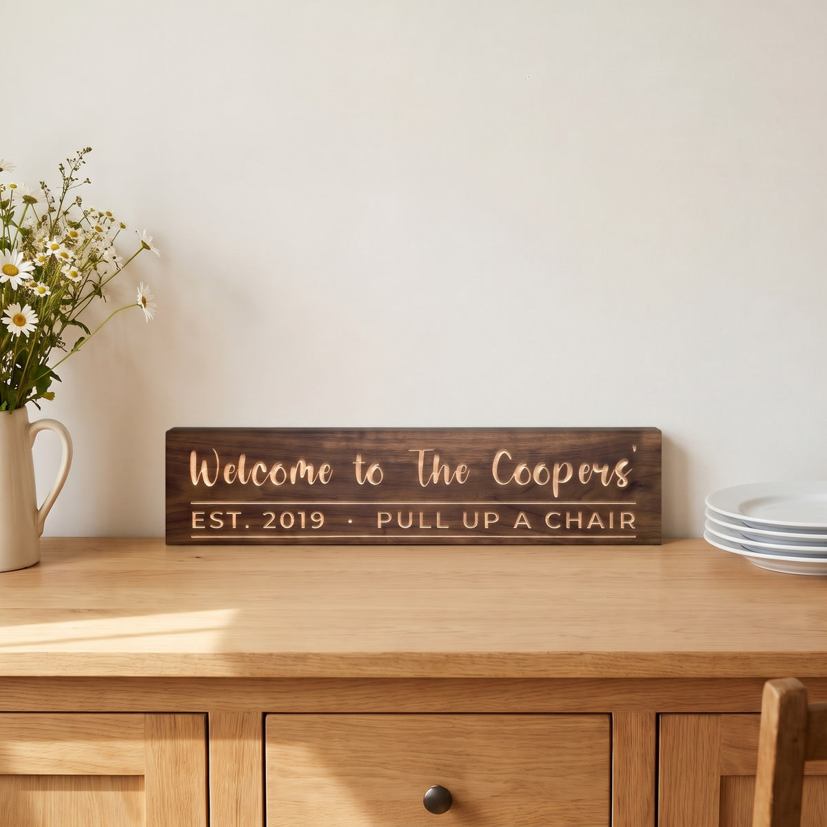 Welcome to The Coopers' personalized wood sign standing on a farmhouse sideboard beside a pitcher of wildflowers and a stack of plates