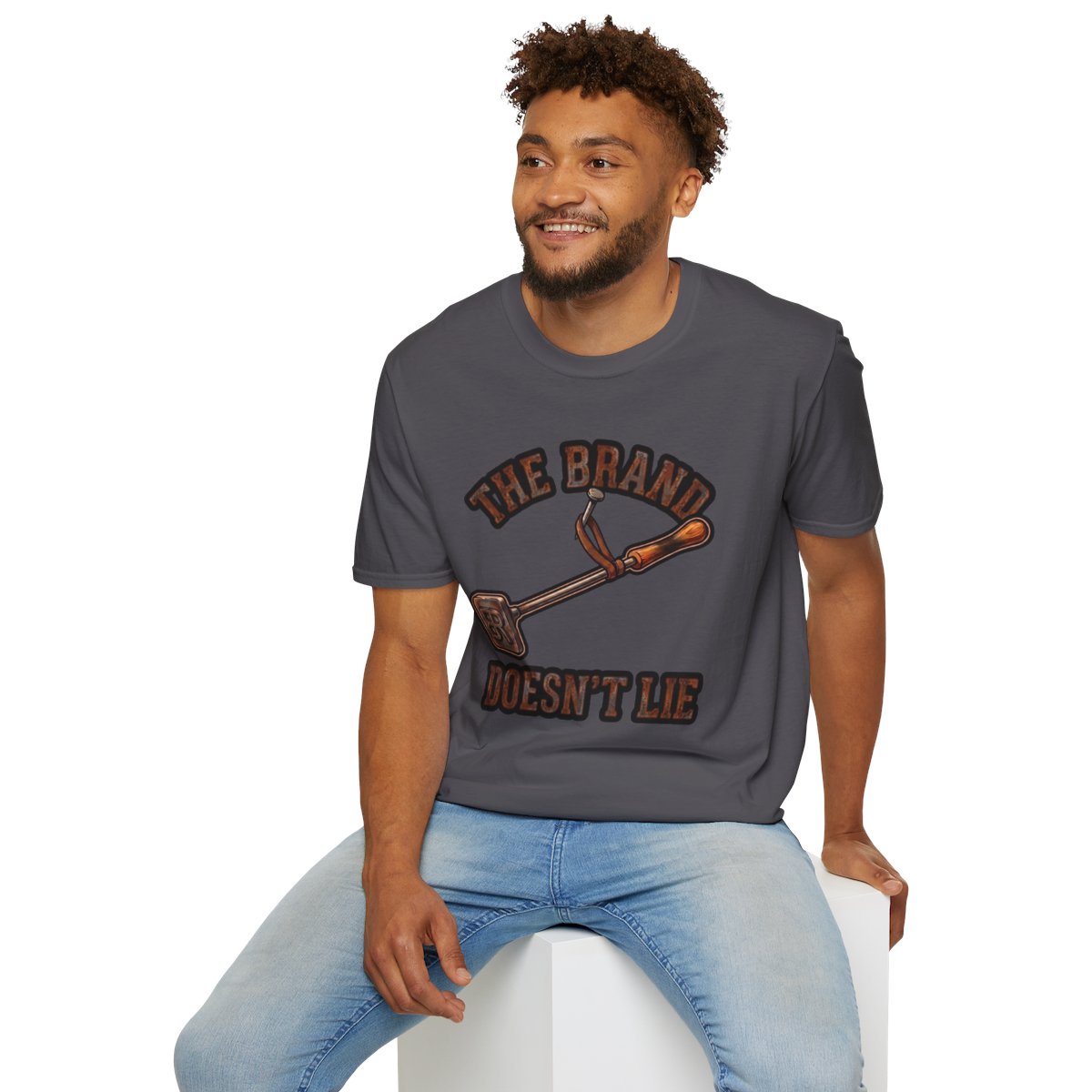 A smiling man in a dark gray Ranch Shirt - The Brand Doesn't Lie, seated on a white block, from Bluegrass Gifts