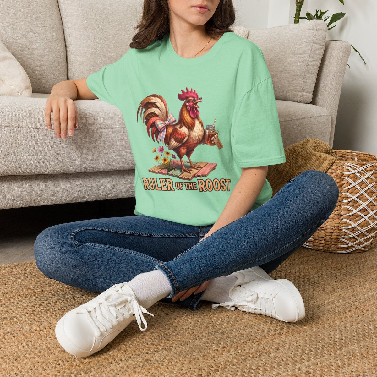 A woman sits on a rug, wearing a light green oversized Rooster Shirt - Ruler of the Roost, with a couch behind her