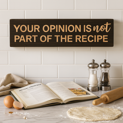 Rustic farmhouse kitchen scene with wooden wall sign “Your Opinion Is Not Part of the Recipe” above cookbook, flour, and rolling pin.
