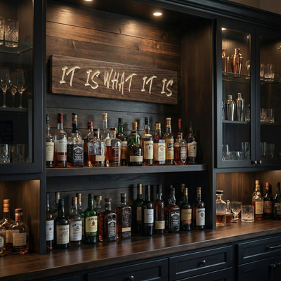 Rustic carved wood sign IT IS WHAT IT IS mounted above shelves of whiskey bottles in a dark wood home bar setting.