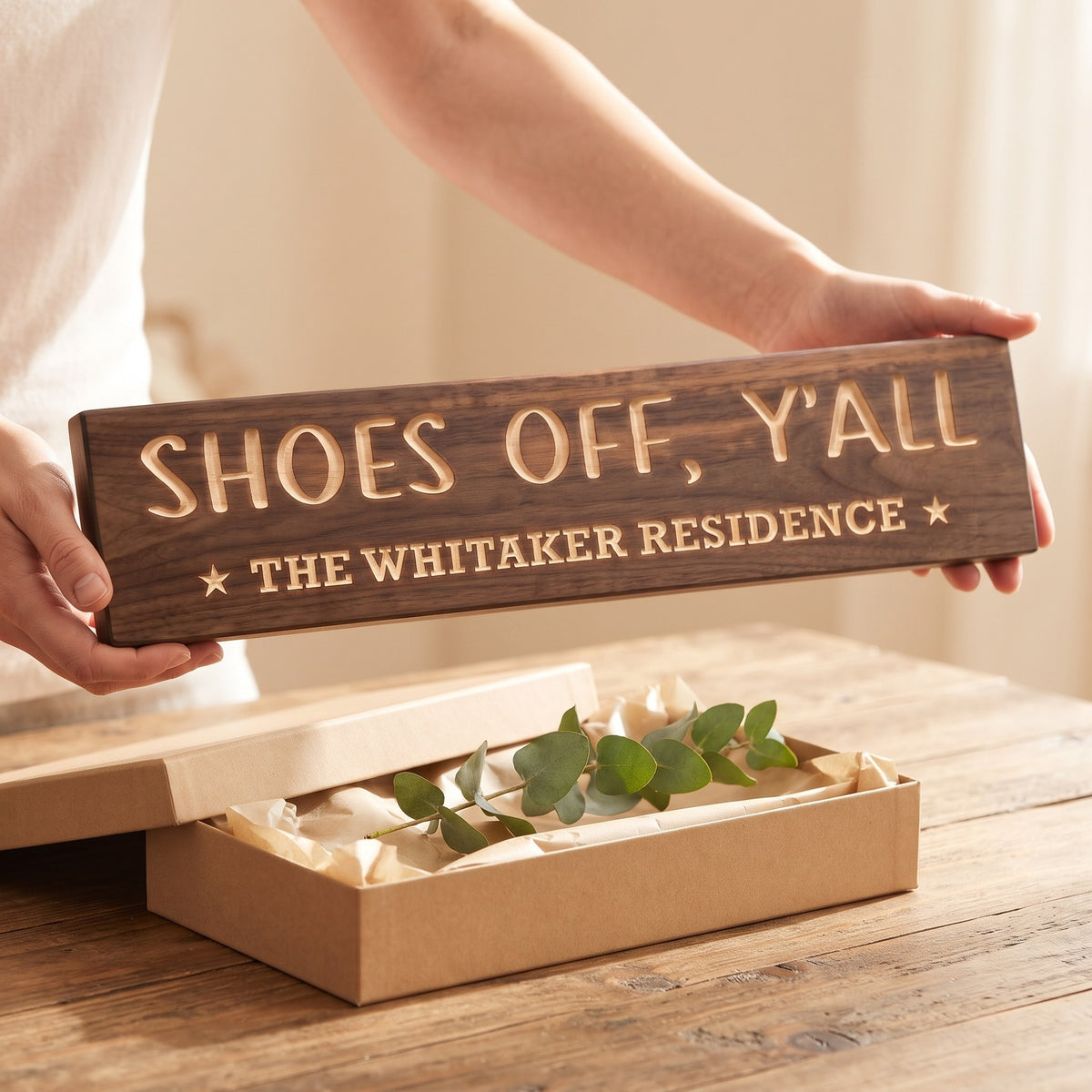 Hands holding a personalized wood sign that reads Shoes Off, Y'all over a kraft gift box with tissue paper and twine