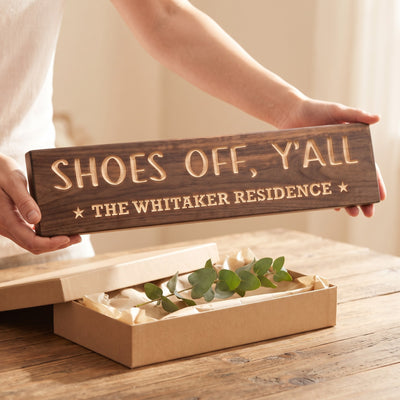 Hands holding a personalized wood sign that reads Shoes Off, Y'all over a kraft gift box with tissue paper and twine