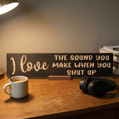 A close-up of the Silence Is Golden Sign on a wooden desk with a coffee mug and headphones, from Bluegrass Gifts
