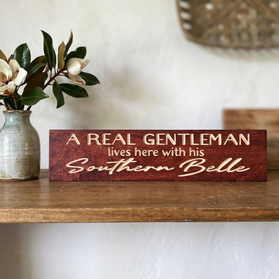 A dark reddish-brown Southern Belle Decor sign with carved letters, on a wooden shelf next to a vase of magnolia from Bluegrass Gifts