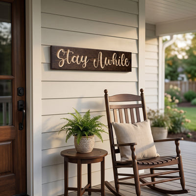 The Stay Awhile sign on a front porch beside the door, above a rocking chair and a potted fern