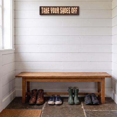 The Take Your Shoes Off sign hanging high on a white shiplap mudroom wall above a wooden bench with shoes and boots underneath