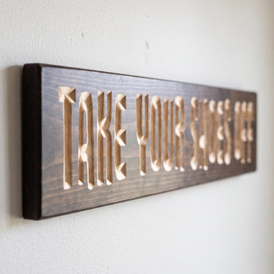 Close-up of the carved letters on the Take Your Shoes Off sign, natural pine inside the cuts against the stained grain