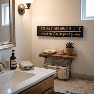Text Me If You Run Out of TP funny bathroom sign hung above a wooden floating shelf with toilet paper basket, candle, and succulent in a modern bathroom