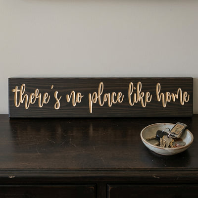 A close-up view of a dark wood There's No Place Like Home Sign resting on a dresser with keys in a bowl, from Bluegrass Gifts