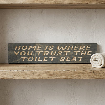 A dark gray Toilet Seat Sign with carved letters rests on a light wooden shelf next to a rolled white towel from Bluegrass Gifts