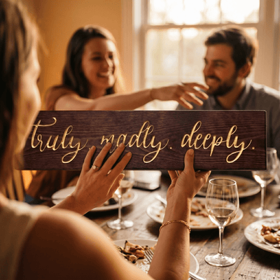Woman with an engagement ring holding the Truly Madly Deeply carved wood sign across a dinner table as friends celebrate together with wine