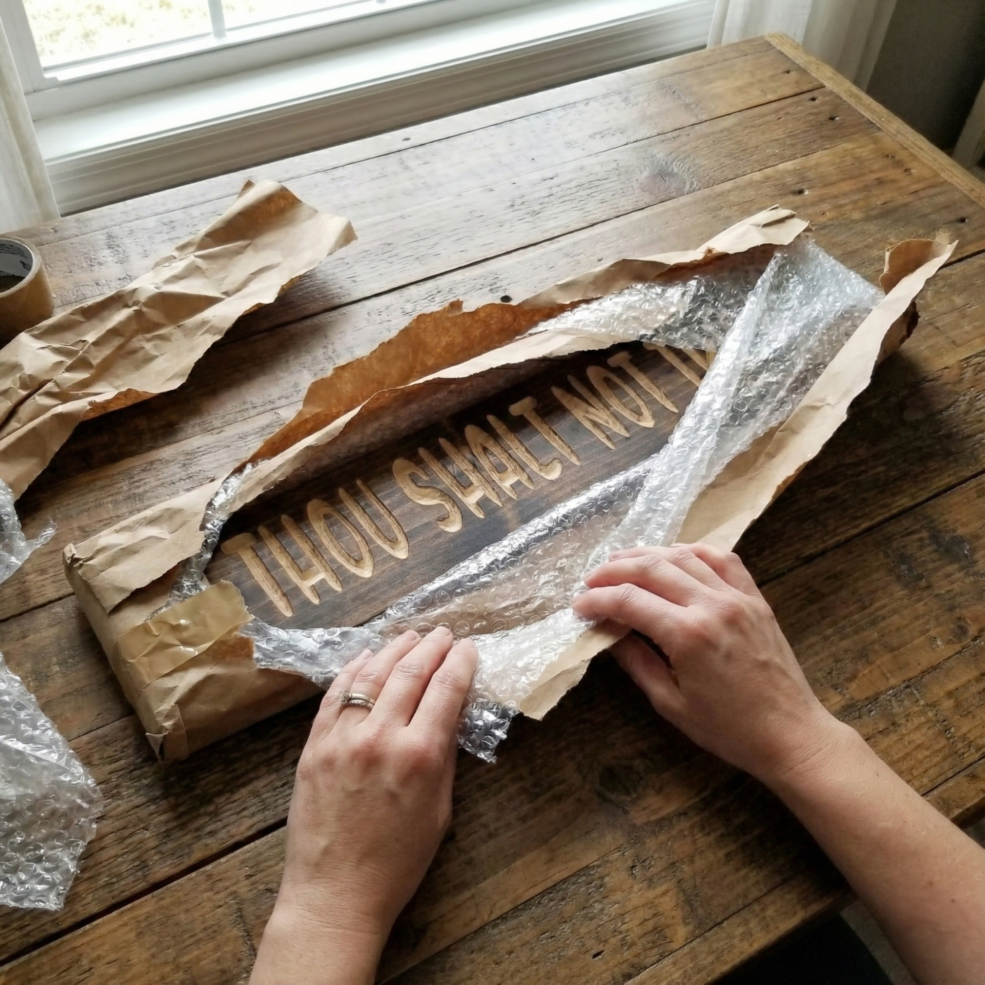 Hands unwrapping a handmade Bluegrass Gifts "Thou Shalt Not Try Me" wooden sign from protective packaging on a rustic wooden table.