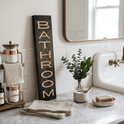 Vertical CNC carved bathroom wood sign styled on a marble counter beside copper fixtures, eucalyptus, and a vintage farmhouse sink — modern farmhouse bathroom decor.