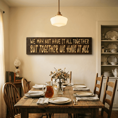 We May Not Have It All Together But Together We Have It All carved wood sign hanging on a cream wall above a rustic farmhouse dining table with wildflowers and a pendant light