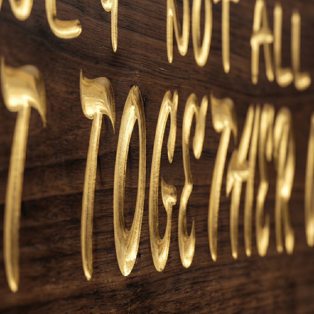 Macro close-up of the deep CNC-carved gold-filled lettering on the We May Not Have It All Together wood sign, showing rich walnut grain and carving depth