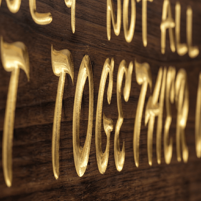 Macro close-up of the deep CNC-carved gold-filled lettering on the We May Not Have It All Together wood sign, showing rich walnut grain and carving depth