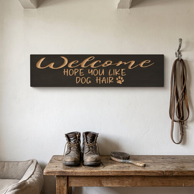The "Welcome Hope You Like Dog Hair" sign is mounted on a white wall above a rustic table with boots and a leash, from Bluegrass Gifts