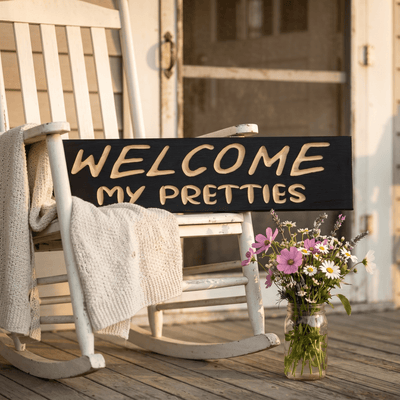 Welcome My Pretties Halloween porch sign resting on a white rocking chair with wildflowers nearby.