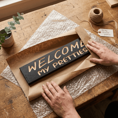 Hands wrapping a Welcome My Pretties wood sign in kraft paper as a gift for a Halloween lover.