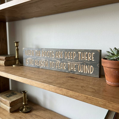 A dark gray When the Roots Are Deep Sign rests on a wooden shelf next to old books and a small potted plant from Bluegrass Gifts
