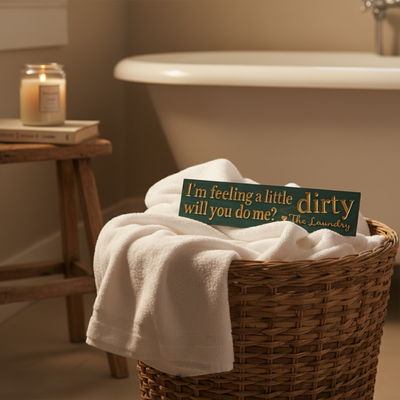 A funny carved wood laundry sign with the quote 'I'm feeling a little dirty' sitting in a wicker laundry basket next to a bathtub.