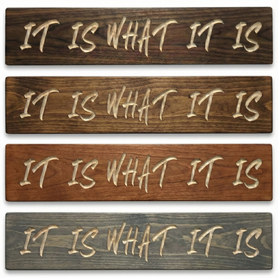 Stack of four carved IT IS WHAT IT IS wood signs showing different finish options including walnut, dark roast, cherry, and weathered gray.