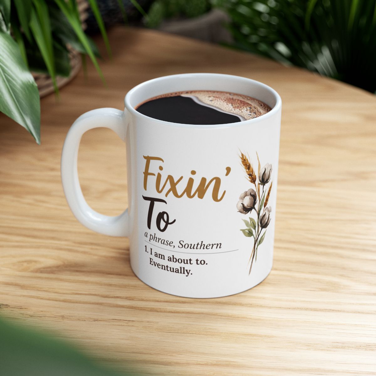 A white ceramic Definition Mug filled with dark coffee, sitting on a warm wooden table with green foliage in the background