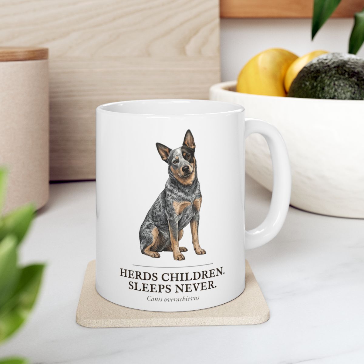 A Dog Mom Mug with a cattle dog design rests on a light coaster on a kitchen counter with fruit and greenery nearby