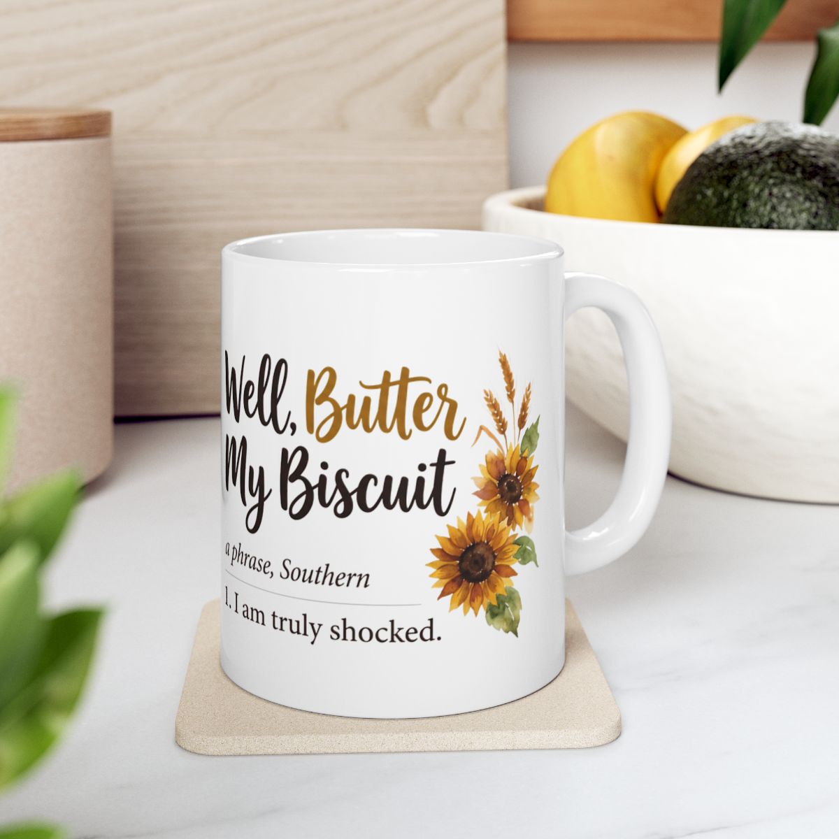 A white ceramic mug with the "Well, Butter My Biscuit" design, placed on a light-colored coaster in a kitchen setting with fruit in a bowl, a practical item among Mugs for Women