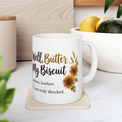 A white ceramic mug with the "Well, Butter My Biscuit" design, placed on a light-colored coaster in a kitchen setting with fruit in a bowl, a practical item among Mugs for Women
