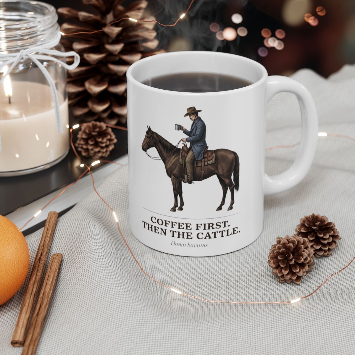 The Cowboy Coffee Mug filled with dark coffee, nestled in a warm holiday setting with pinecones, a candle, and string lights