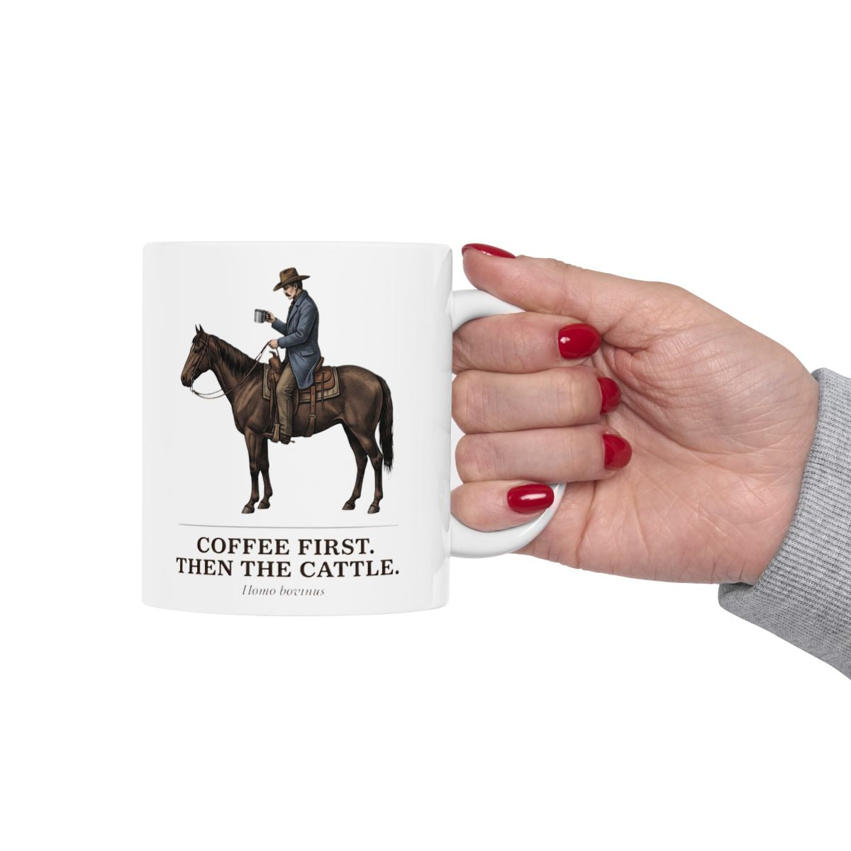 A hand with red nail polish holds the white Cowboy Coffee Mug by its handle against a bright, clean background