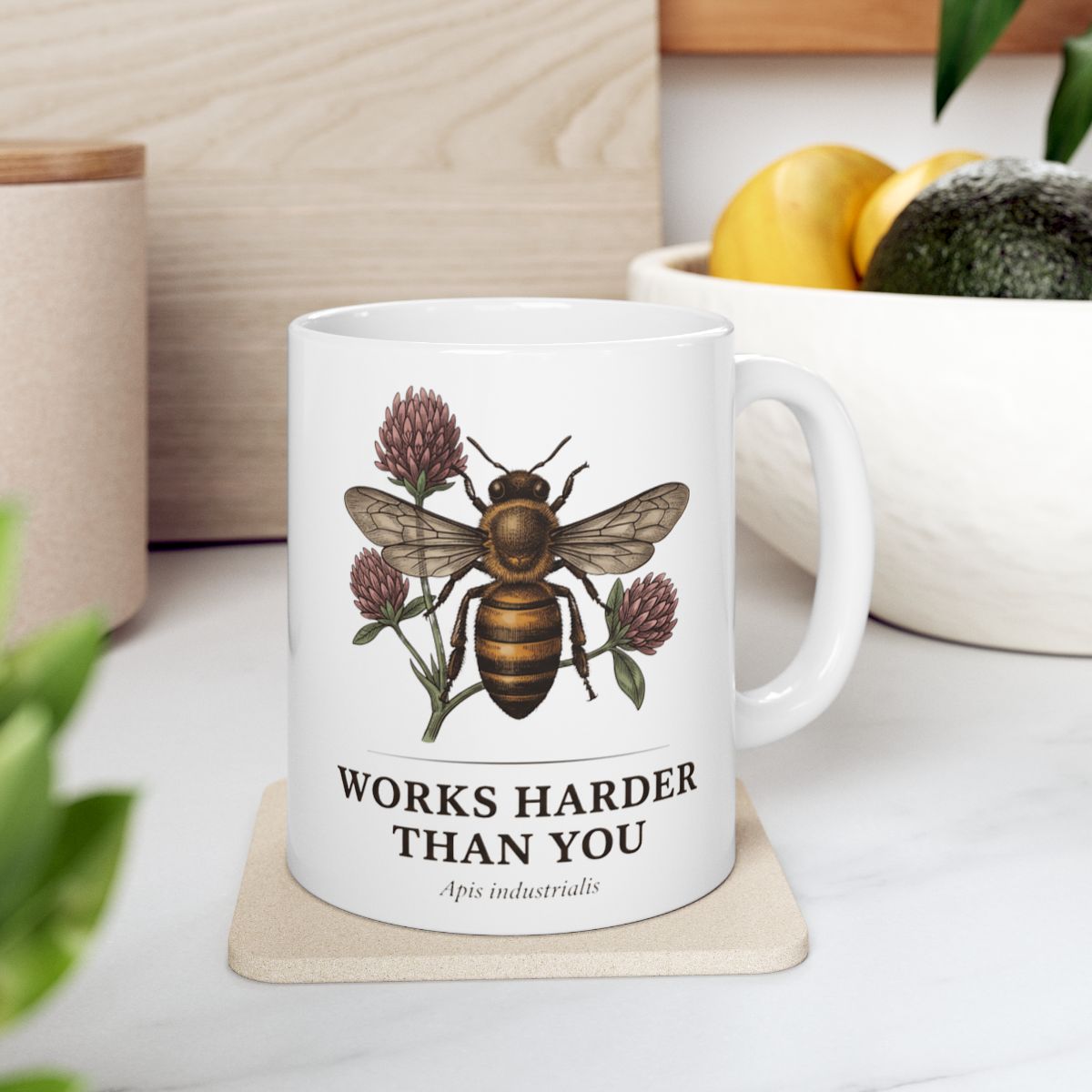 A white Bee Coffee Mug resting on a light-colored coaster, with kitchen items like fruit and an avocado blurred in the background