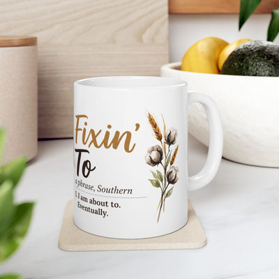 A white ceramic Definition Mug with the "Fixin' To" design, resting on a light coaster in a bright kitchen setting