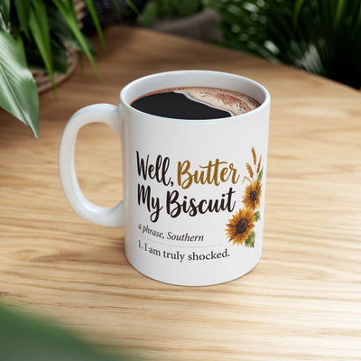 A white ceramic mug filled with coffee, displaying the "Well, Butter My Biscuit" design, sitting on a wooden table with green foliage in the background, a welcoming option for Mugs for Women