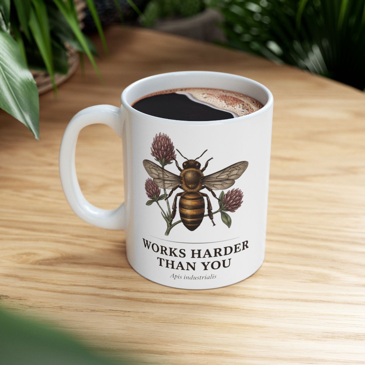 A white Bee Coffee Mug with coffee, placed on a warm wooden table, with green foliage subtly visible in the background