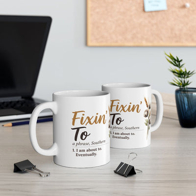 Two white ceramic Definition Mugs with the "Fixin' To" design, one in front, on a desk with a laptop and plant