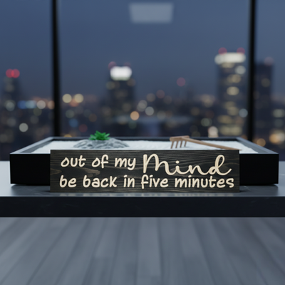 Humorous wooden desk sign "be back in five minutes" placed in front of a desktop Zen garden with a city skyline at night.